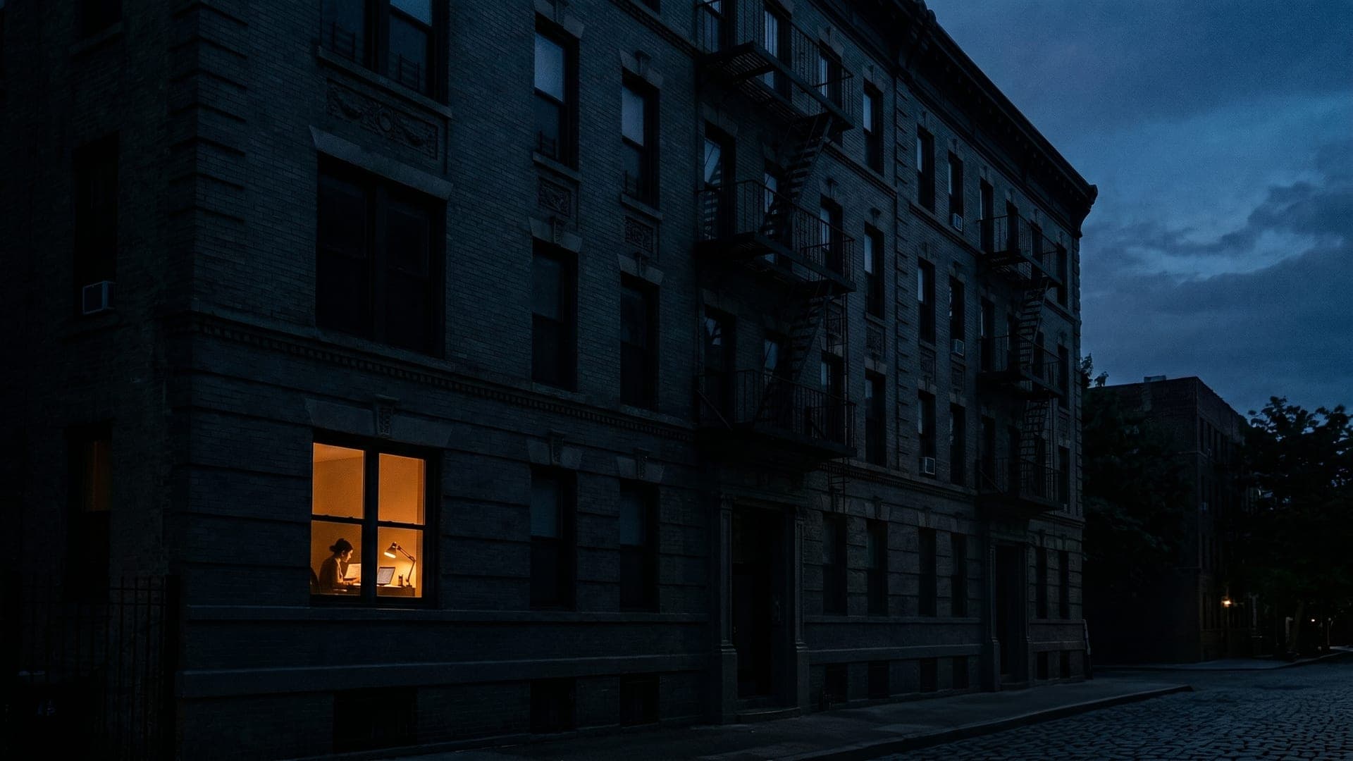 A single lit window glowing in a dark building at dusk — someone still up, still applying.